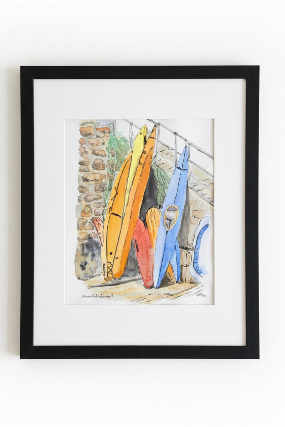 A framed watercolor painting of colorful kayaks leaning against a stone wall, presented in a black frame with a white mount