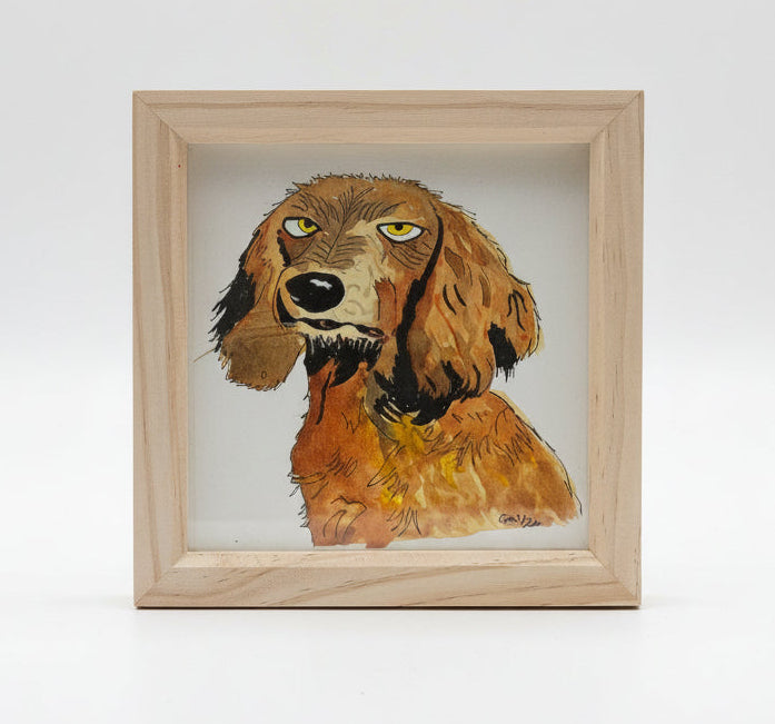 A framed watercolor painting of a brown and golden retriever dog with yellow eyes, set in a light natural wood frame and displayed on a wooden surface.