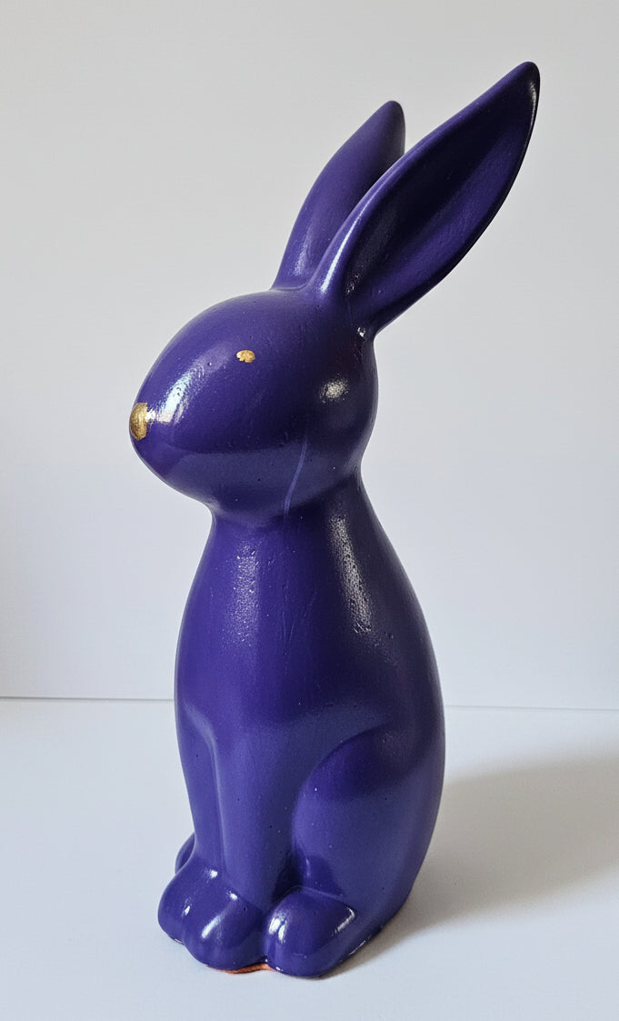 A glossy purple rabbit figurine with small gold accents on its nose and eyes, standing on a white surface against a white background.